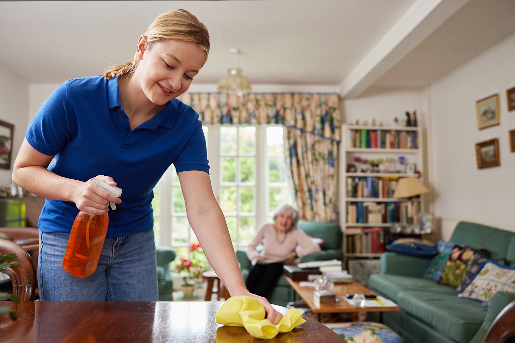 Female Home Help Cleaning House And Talking To Senior Woman
